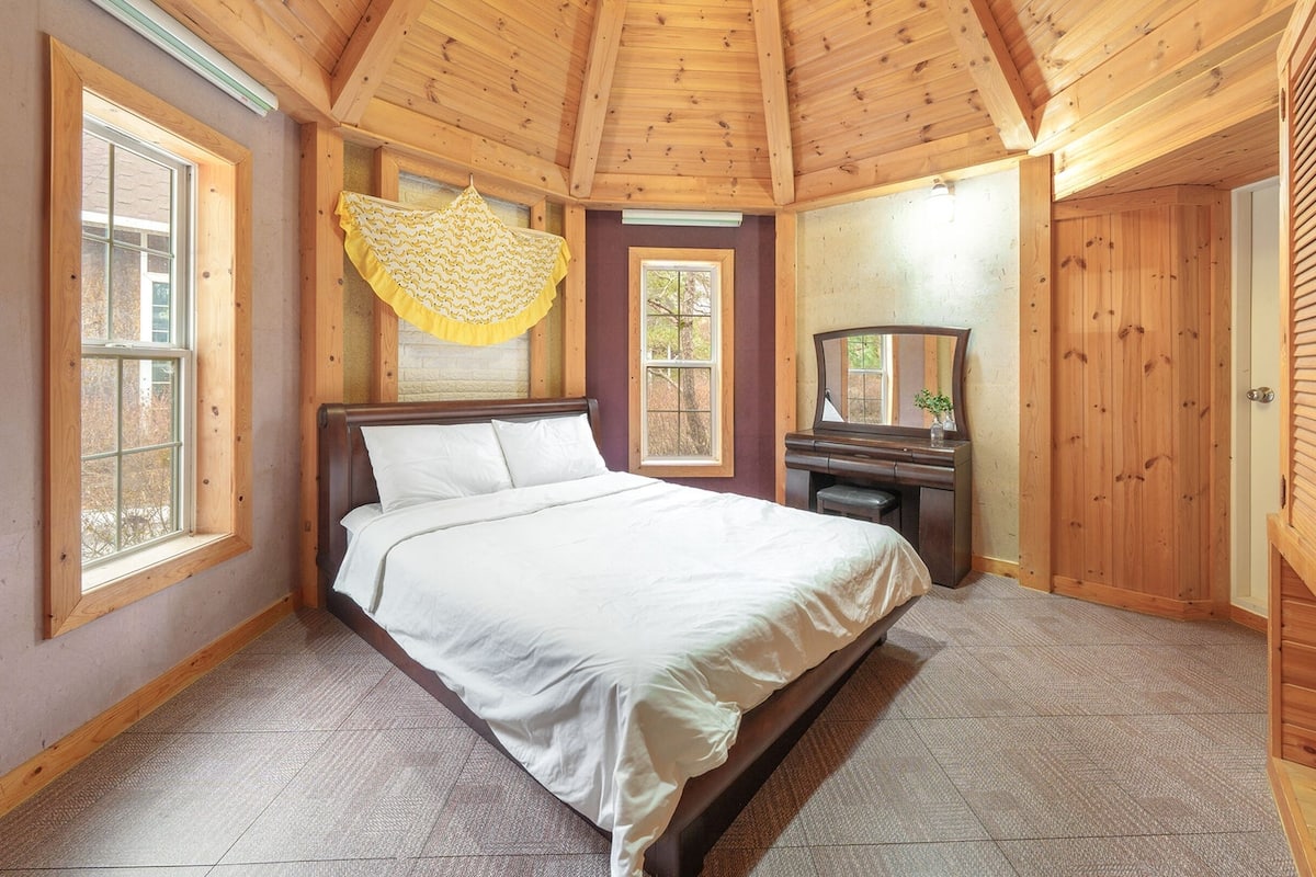 Ein Schlafzimmer mit Holzwänden, einem Bett, zwei Fenstern, einem Schminktisch und einem Spiegel.
