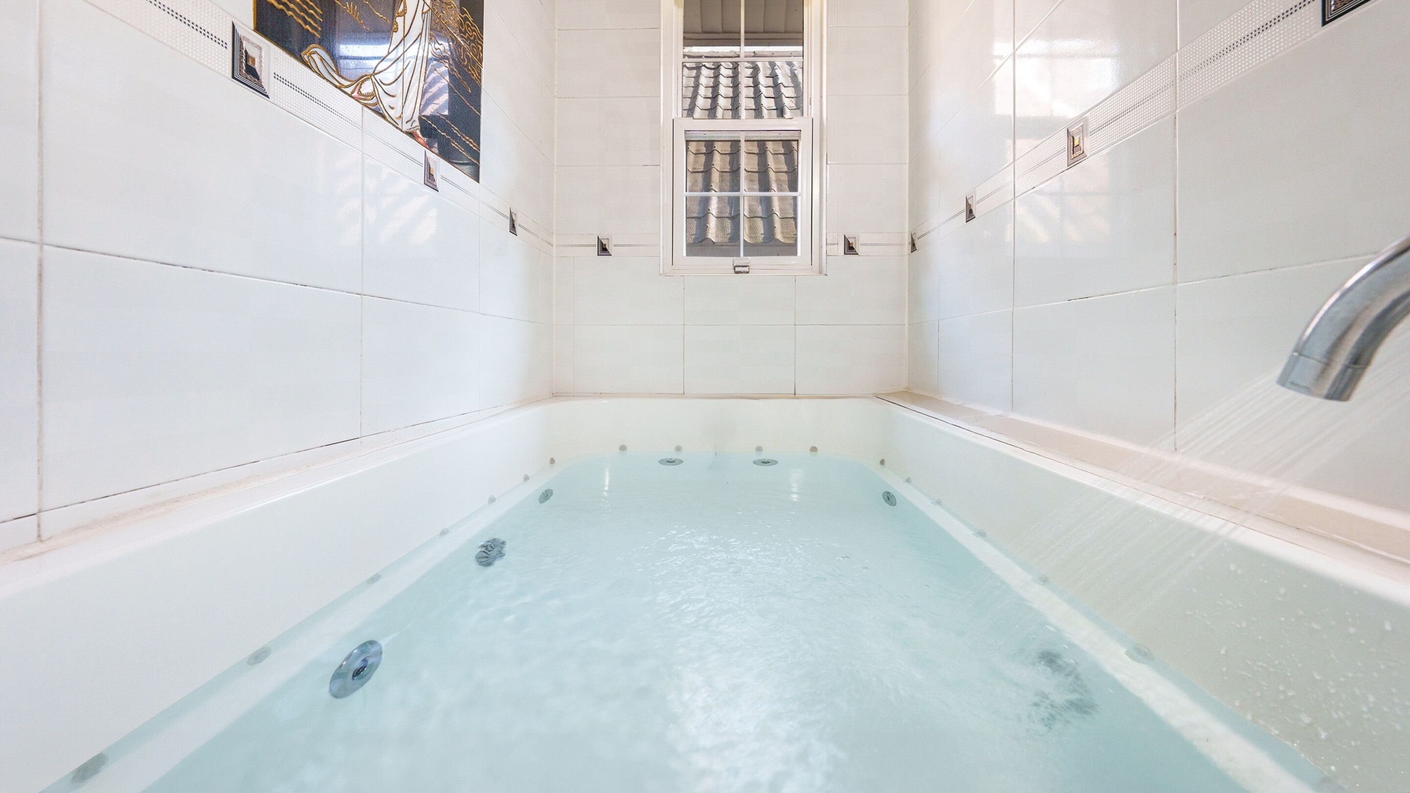 Eine große, leere Badewanne mit Fenster darüber, umgeben von weißen Fliesen.