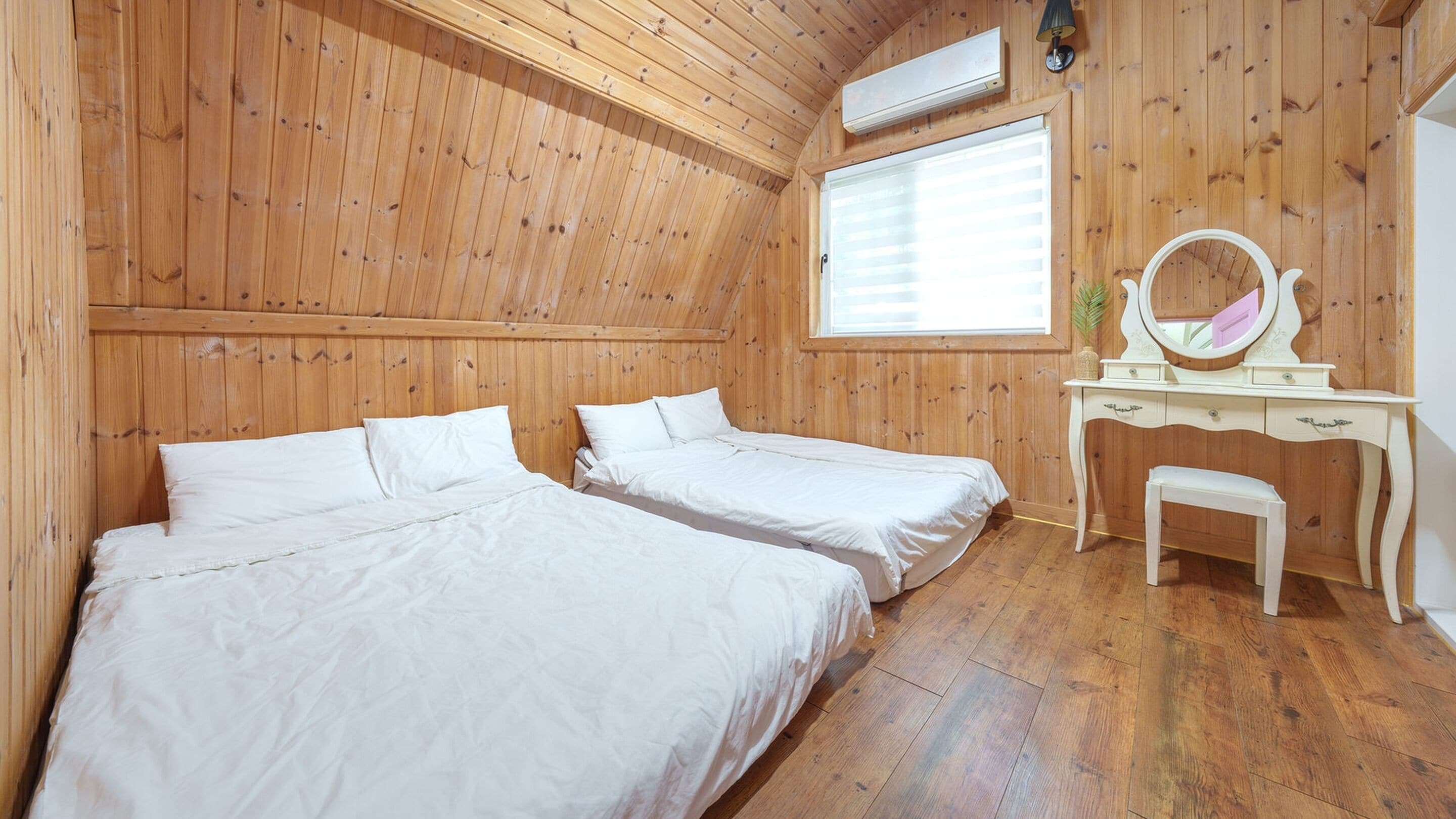 A room with wooden walls and floors, two beds with white bedding, a wooden dresser with a mirror, and a window with blinds.