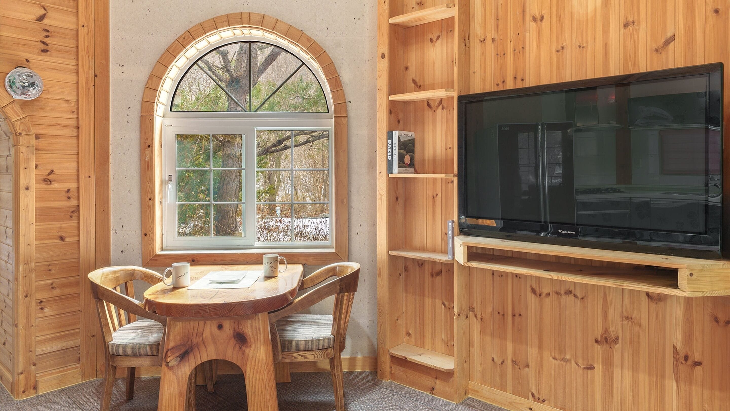 Una habitación acogedora de madera con un amplio ventanal arqueado, un mueble de televisión de madera y una pequeña mesa con sillas.