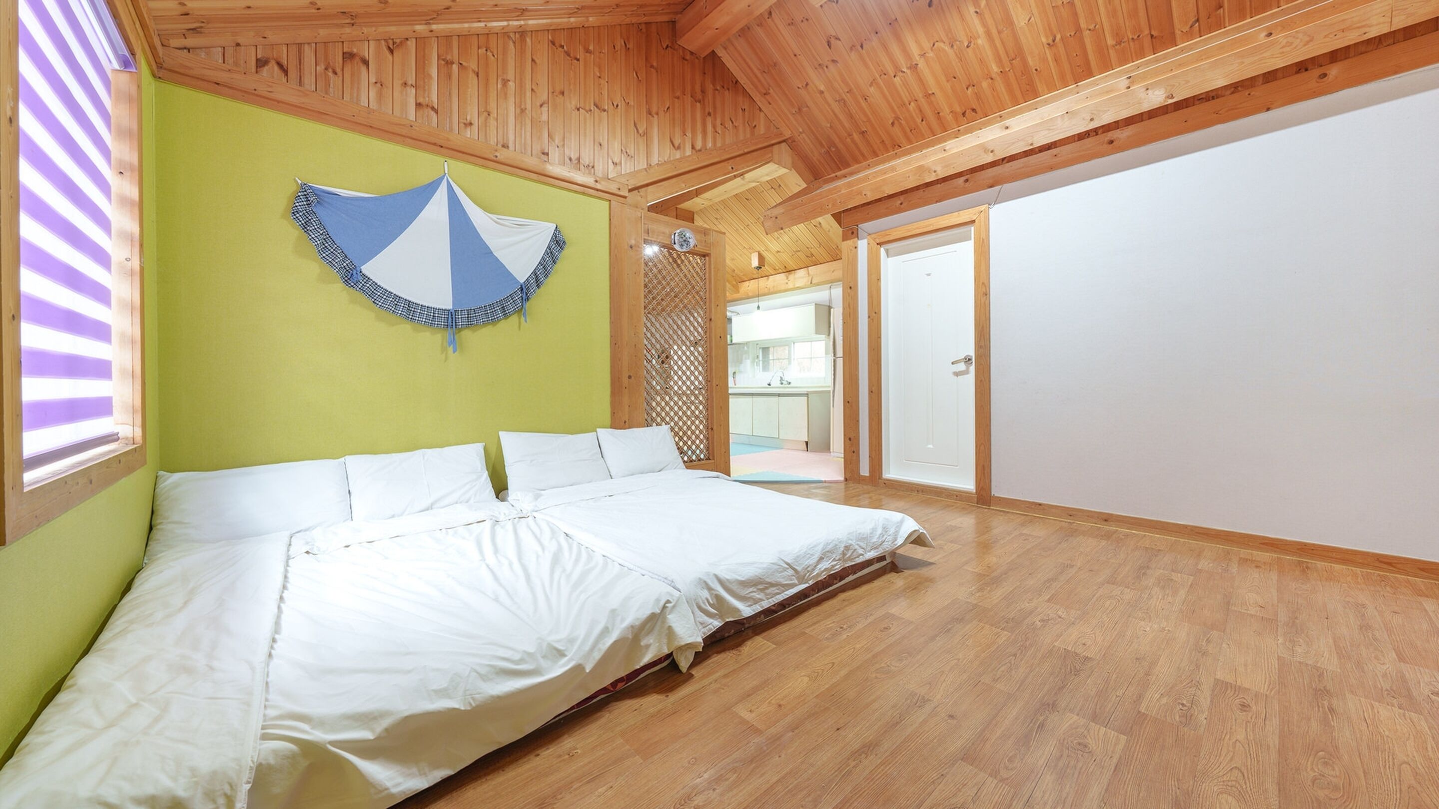 Un grand lit dans une chambre dont le plafond est orné de poutres en bois, un mur est peint en vert et d’où l’on aperçoit un coin cuisine.