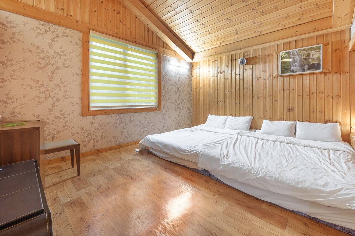 Ein Schlafzimmer mit Holzboden, einem Bett mit weißer Bettwäsche, einem Schreibtisch und einem Fenster mit Jalousien.