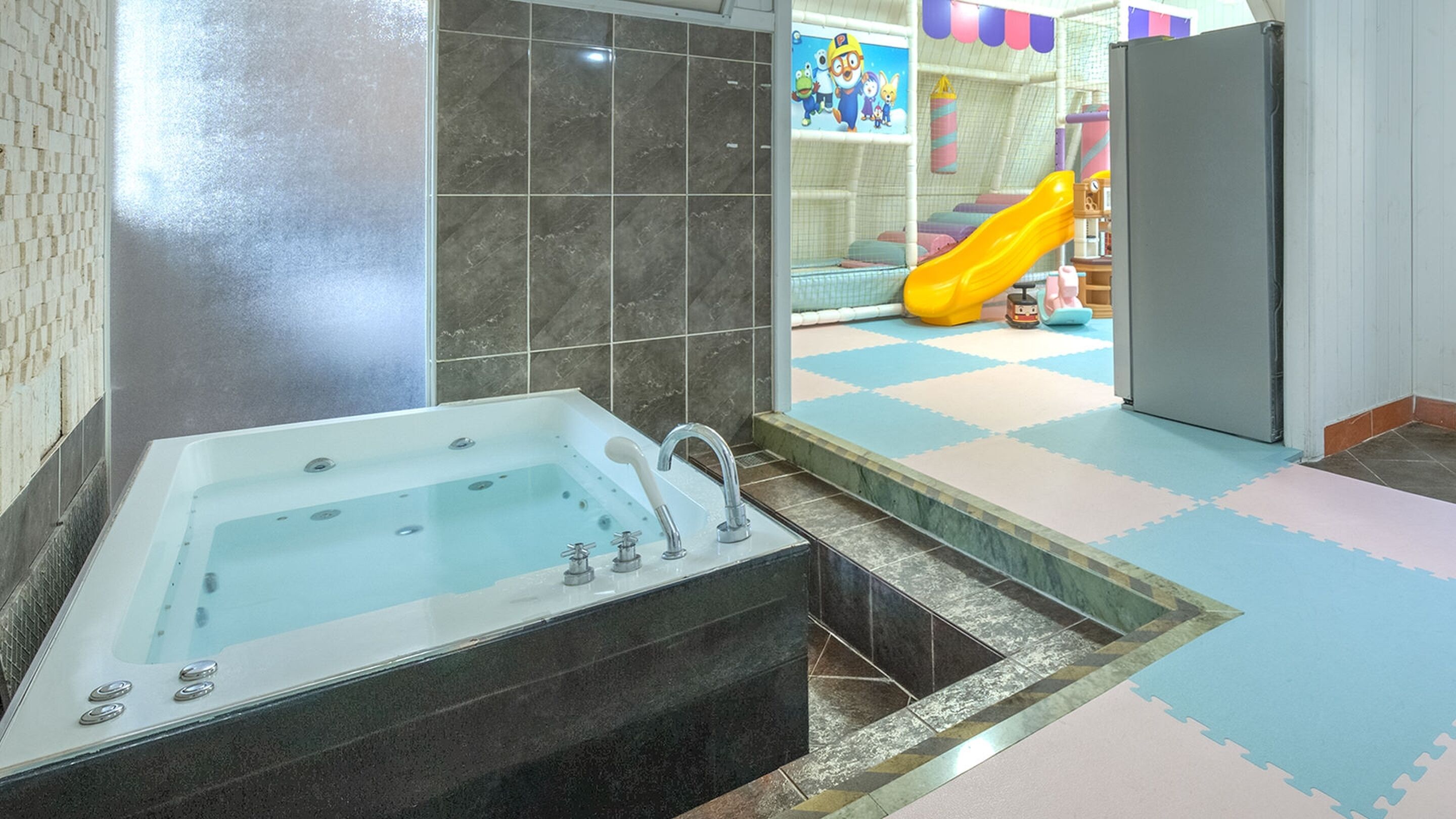 Un baño con una bañera grande, una zona de ducha y un área de juegos para niños visible a través de un panel de vidrio.