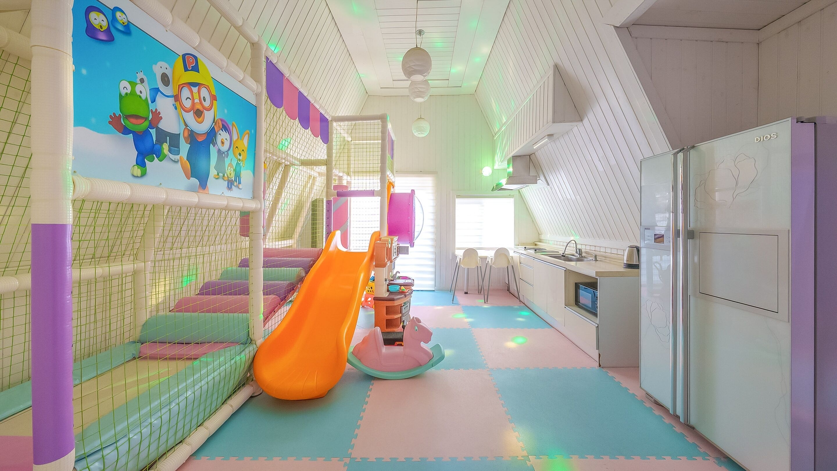 Zona de juegos infantiles con un tobogán, alfombras de juego coloridas y una zona de cocina con refrigerador.