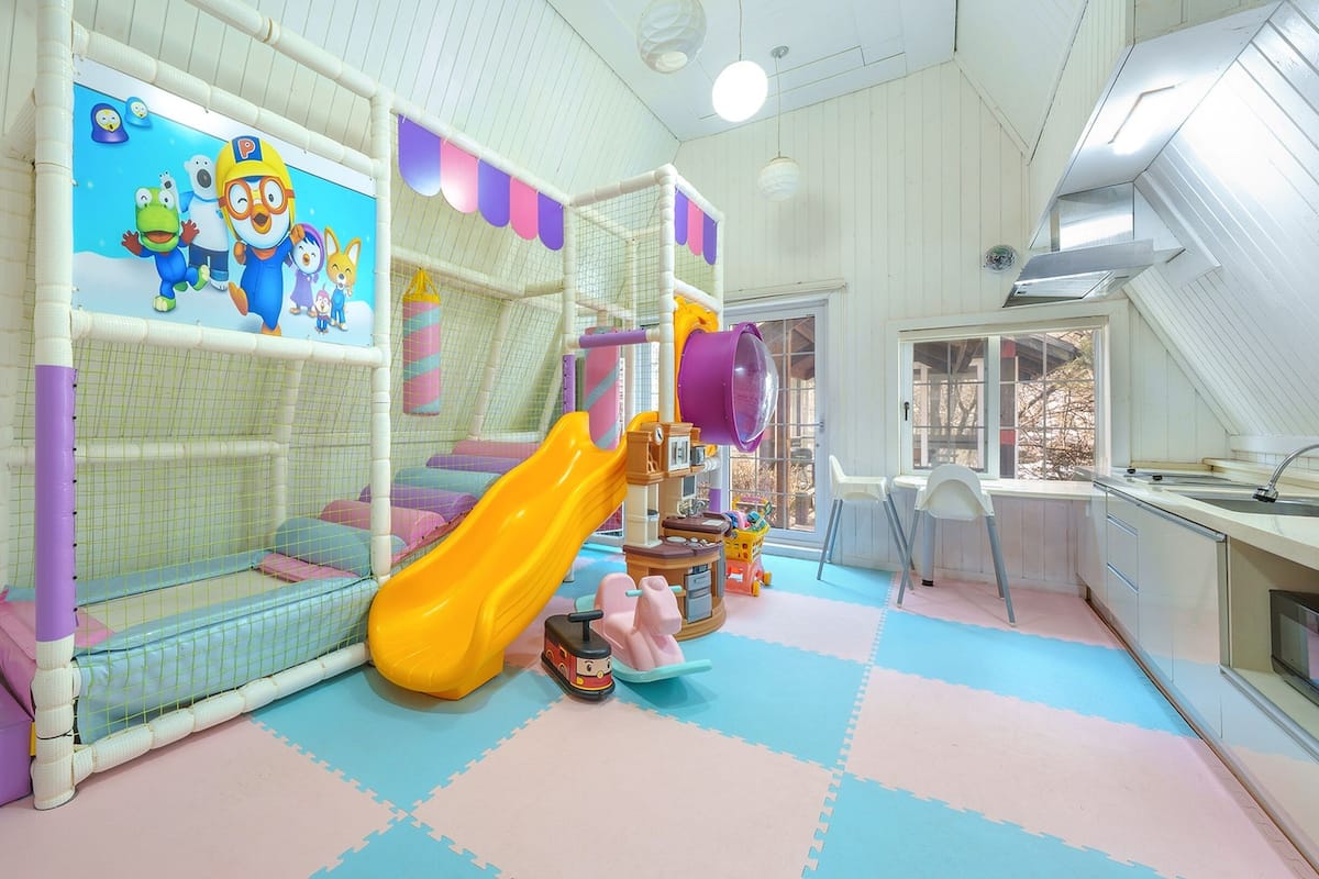 Ein Kinderspielbereich mit Rutsche, Spielgeräten und einem Fenster mit Blick auf Bäume.