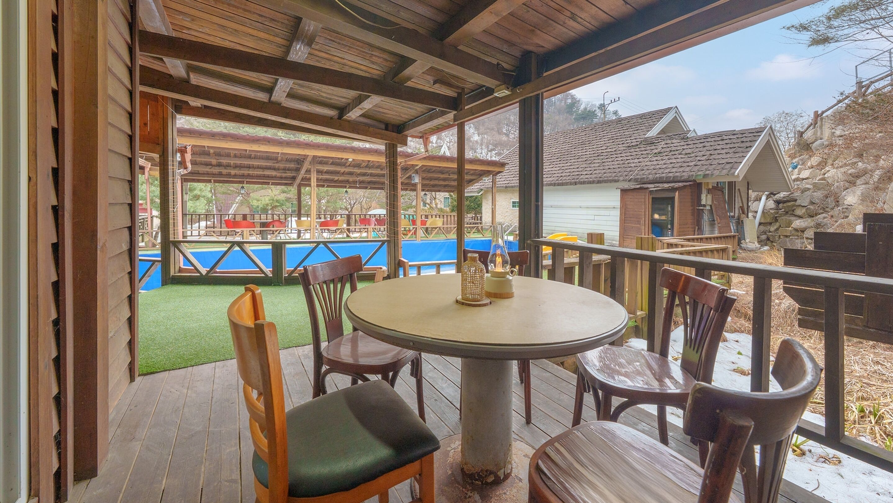 A covered outdoor seating area with a round table and chairs, overlooking a pool and a playground.