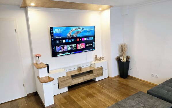 A modern living room with a mounted TV displaying various channels, a wooden console table with a vase and a decorative item, and a potted plant. - Stylish 80 sqm apartment in the heart of the Ruhr area (Bochum)