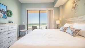 A bedroom with a large bed, a dresser, a chair, and a view of the beach through a sliding door.