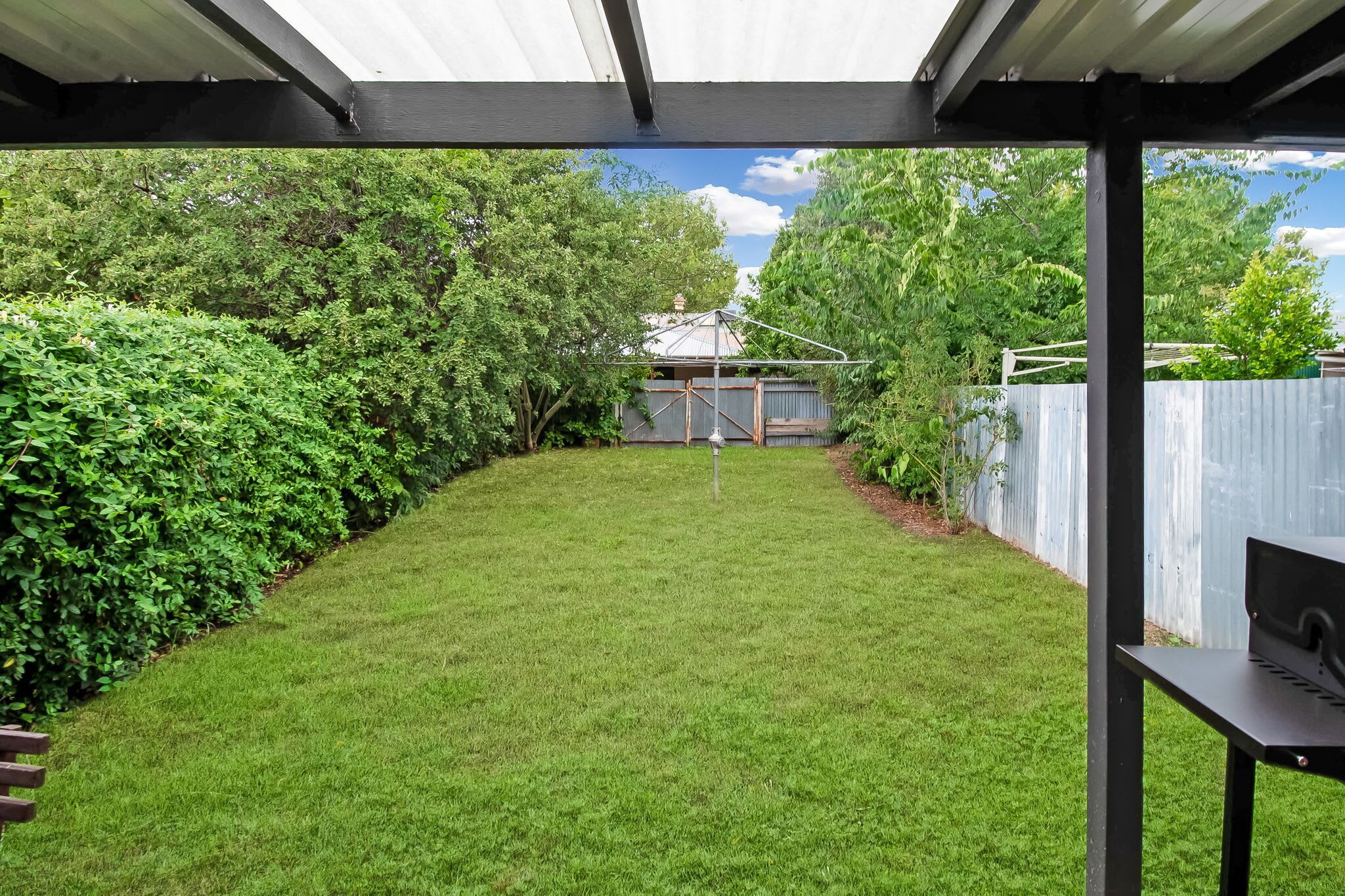 A covered patio area with a grassy lawn, a wooden bench, and a view of a house and trees.