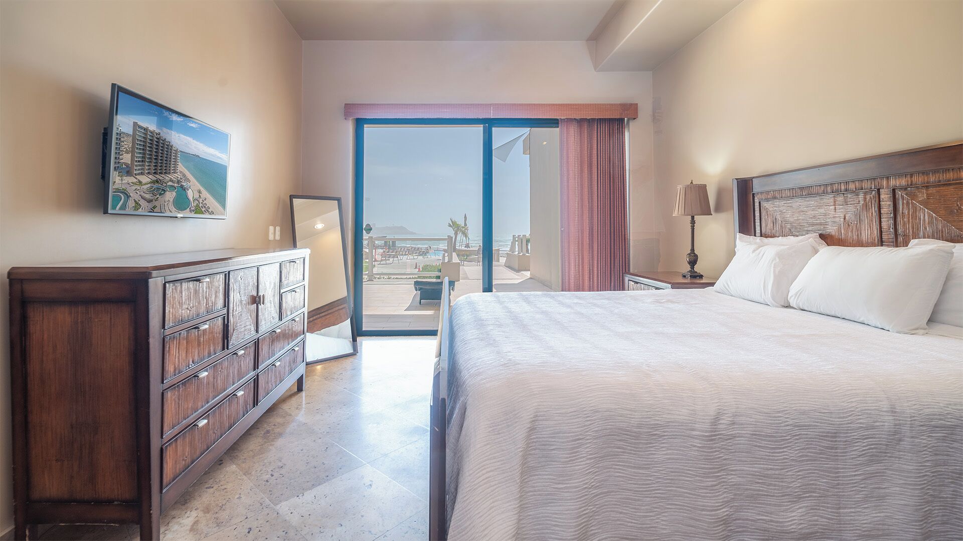 Une chambre d’hôtel avec un lit, une commode, un tableau, une lampe et une vue sur la plage et l’océan, visible par une porte coulissante.