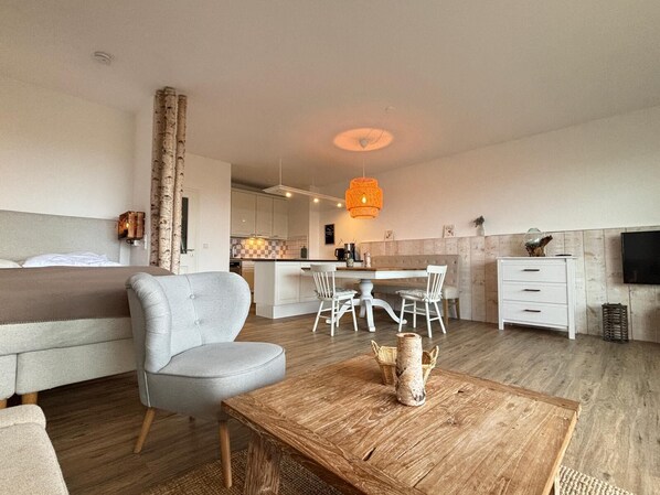A modern hotel room with a bed, a grey armchair, a wooden coffee table, a dining area with a table and chairs, a kitchen in the background, and a TV on the wall. - Stuuv 8 - Vacation in the 1st row - with Baltic Sea view and direct beach location (Scharbeutz)