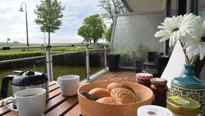 Ein Tisch mit Frühstücksgebäck, einer Kaffeekanne und Gläsern, auf einem Balkon mit Blick auf eine Hecke und Bäume.