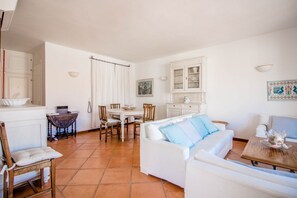 Una sala de estar con un sofá blanco, una mesa de comedor con sillas, una zona de cocina con armarios y un suelo de baldosas.