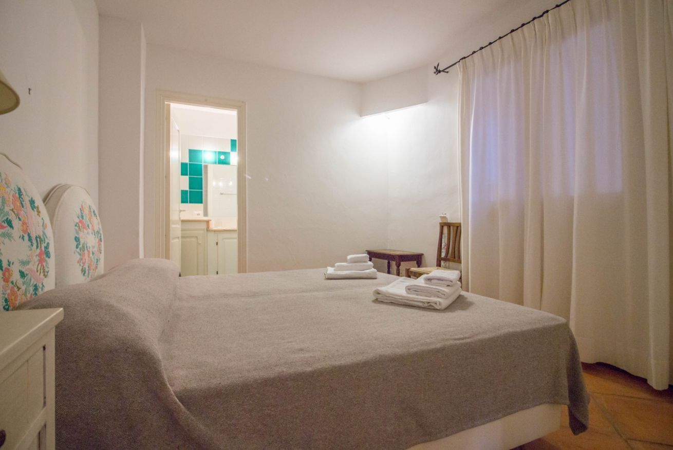 A bedroom with a bed, a small table, a chair, and a bathroom visible through an open door.