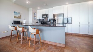 Una cocina moderna con armarios blancos, una isla central con taburetes de bar, electrodomésticos de acero inoxidable y piso de baldosas.