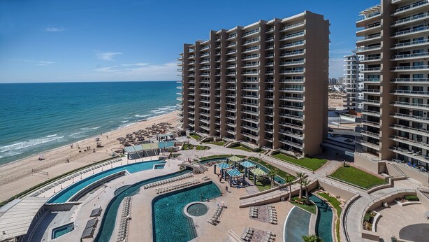 Un hotel de altura con varios balcones, un área de piscina con tumbonas y vista al mar.