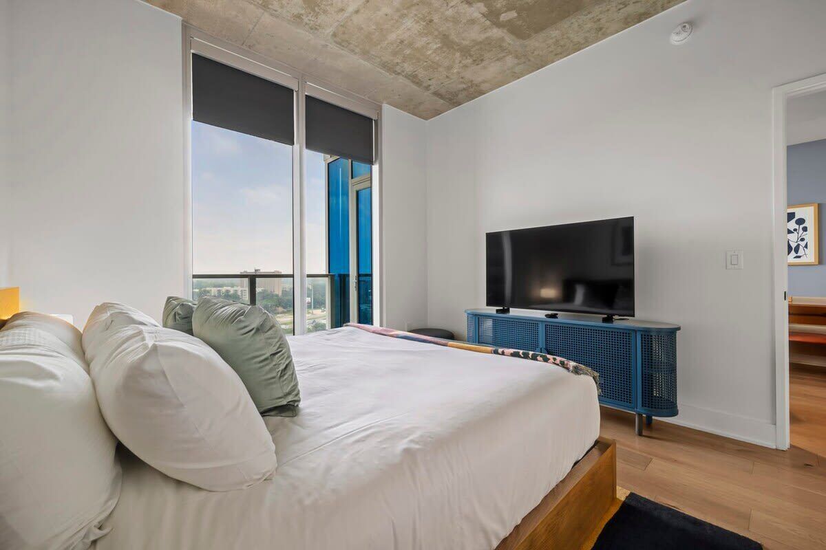 Ein Schlafzimmer mit einem großen Bett, einem Fernseher auf einem blauen Schrank und Blick auf die Stadt durch ein großes Fenster.