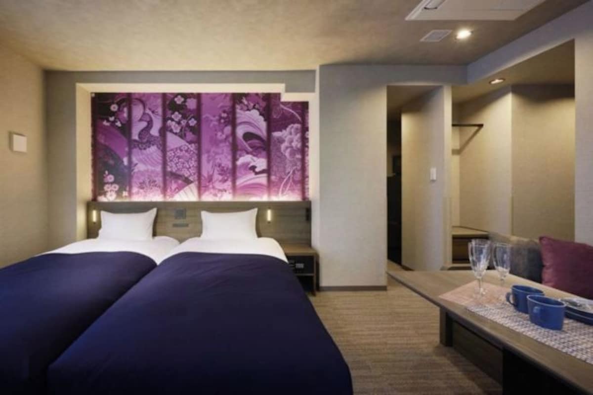 Habitación de hotel con dos camas, un cuadro abstracto grande en la pared, una mesita con un florero y dos vasos, y un banco con cojín.