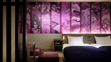Una habitación de hotel con cama, mesita de noche y mesita auxiliar con reloj digital.