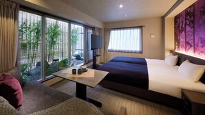 A hotel room with two beds, a dining table, a TV, and large windows offering a view of greenery.