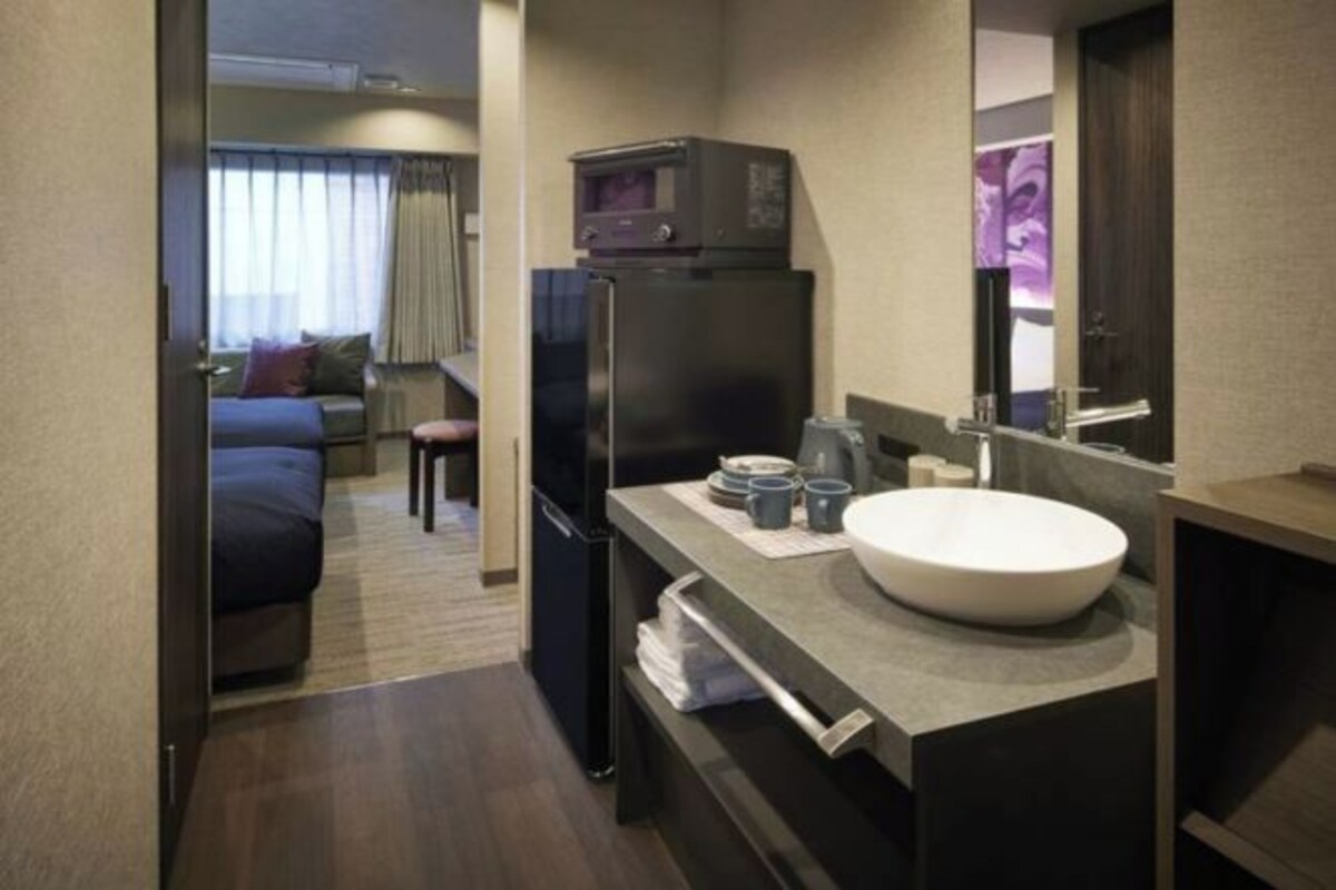 Una habitación de hotel compacta con kitchenette, una cama, zona de estar y baño con lavamanos y espejo.