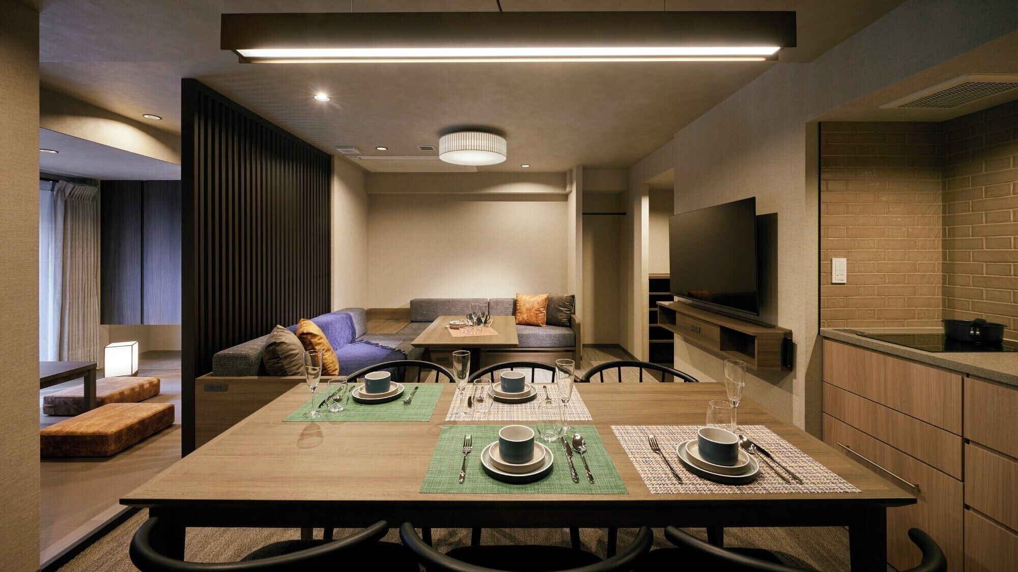 A modern hotel room with a dining area, sofa, TV, and kitchenette.