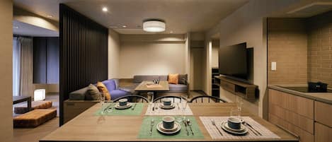 A modern hotel room with a dining area, sofa, TV, and kitchenette.