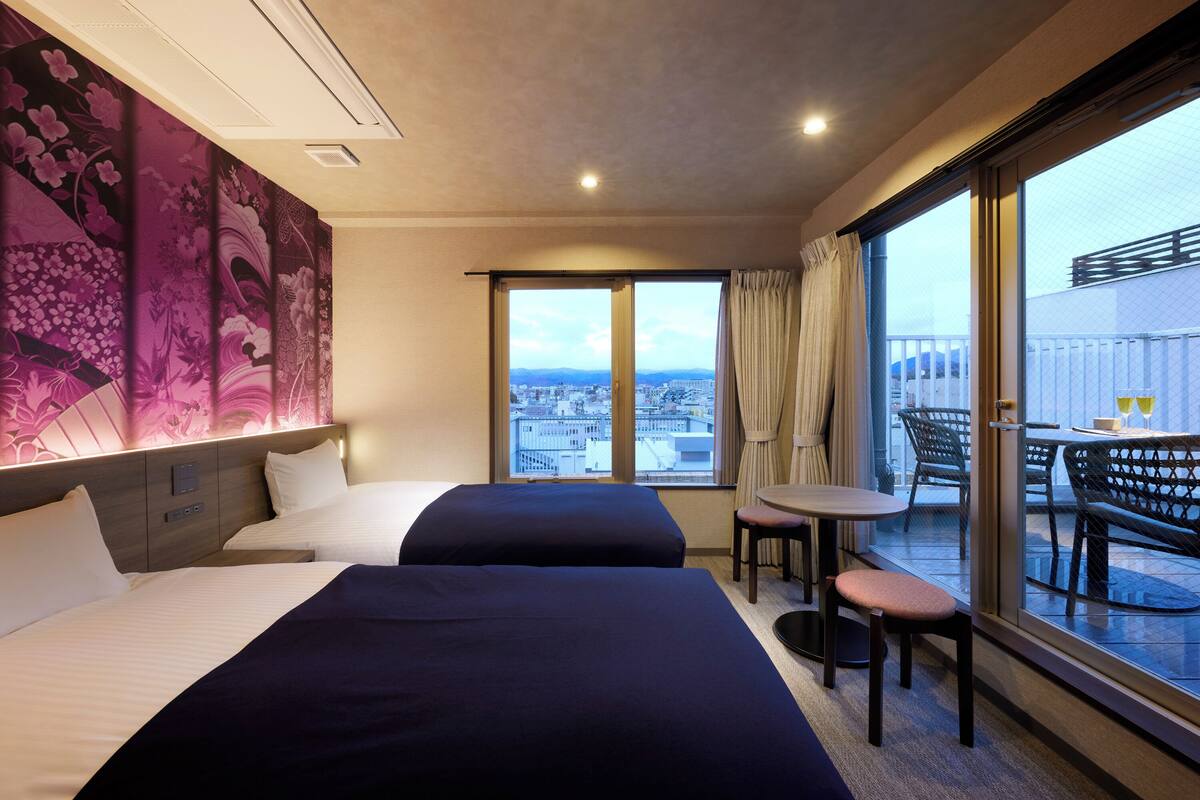 Een hotelkamer met twee bedden, een kleine tafel, een stoel en een balkon met uitzicht op de stad.