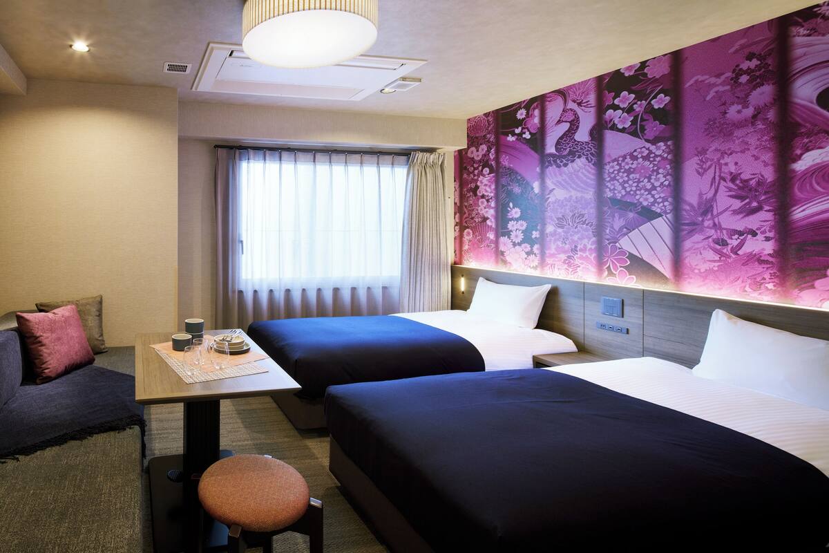 Een hotelkamer met twee bedden, een tafel met een placemat, een stoel, een bank en een groot bloementapijt aan de muur.