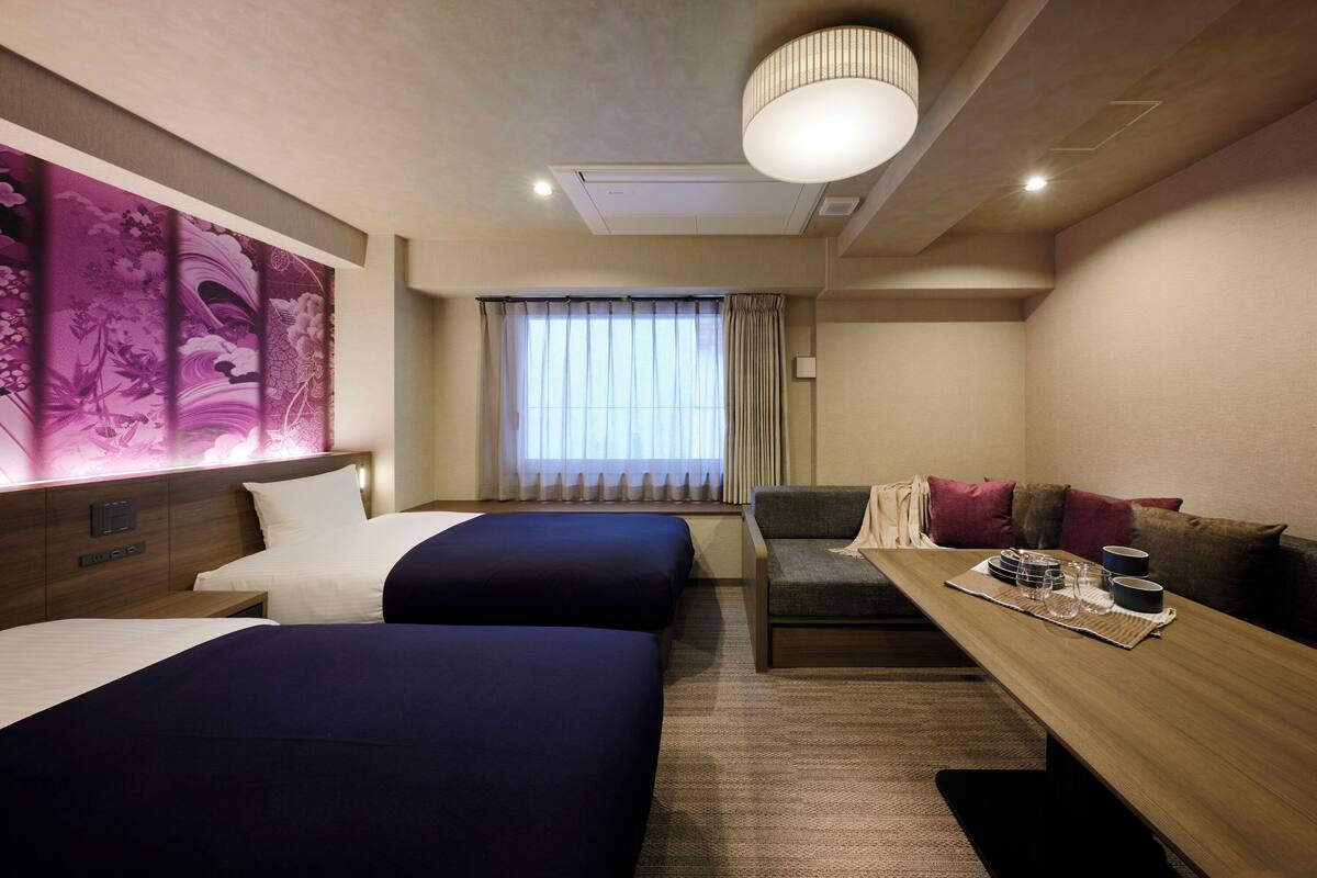 Een hotelkamer met twee bedden, een bank, een eettafel met bekers en een glas, en een groot raam met gordijnen.