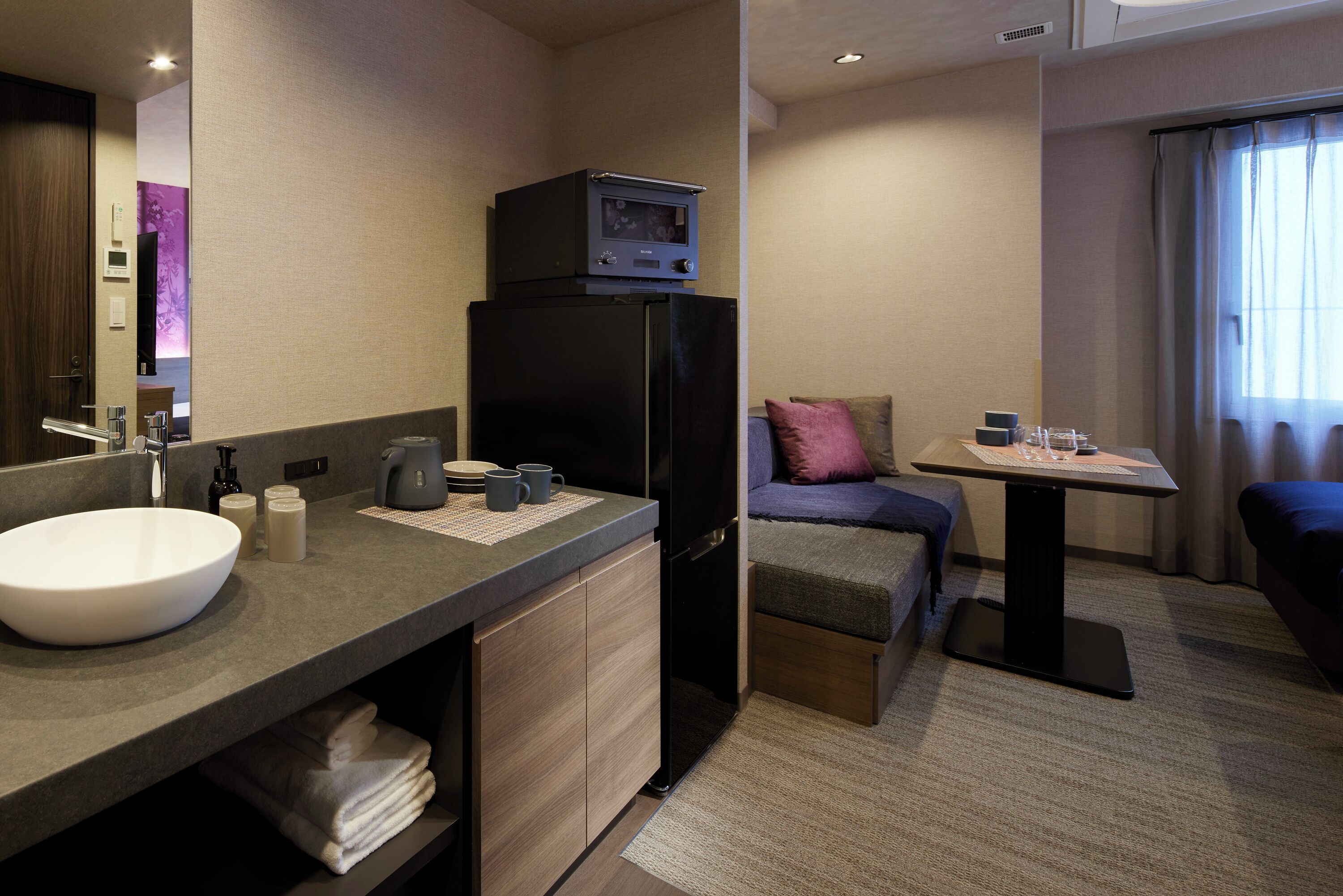 Et kompakt hotelværelse med et badeværelse, der indeholder en vask, et spejl og et sort køleskab. Der er et lille bord med en kop og en fjernbetjening, samt en sofa med en pude.