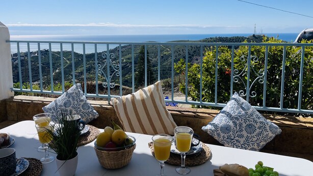 Mesa posta com itens de café da manhã e copos de suco de laranja, com vista para a paisagem costeira.
