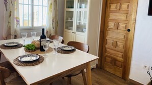 Uma área de refeições com uma mesa posta para dois, incluindo uma garrafa de vinho, copos e pratos. É visível um armário com portas de vidro ao fundo.