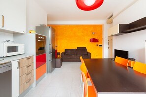 A compact kitchen with a microwave, oven, and refrigerator, a dining table with orange chairs, and a sofa in the background.