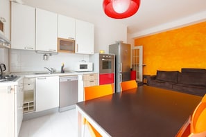 A modern kitchen with white cabinets, a microwave, an oven, and a dishwasher. There is a dining area with a brown table and orange chairs.