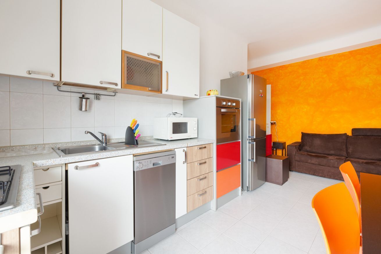 Ett modernt kök med vita skåp, en mikrovågsugn och en diskmaskin. I vardagsrummet finns en röd och orange soffa.