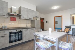 Une cuisine moderne avec des meubles gris, un four encastré et un coin repas comprenant une table blanche et des chaises transparentes.