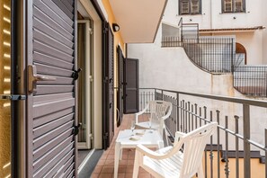 Un balcon avec des chaises blanches et une table, une porte coulissante fermée et un escalier menant à l’étage supérieur.
