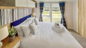 Ein ordentlich bezogenes Bett mit Kopfteil, ein Nachttisch mit Lampe und Pflanze, und ein Blick ins Freie durch ein Fenster.
