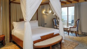 A four-poster bed with white bedding, a wooden headboard, and a canopy. There is a small bedside table and a chair with a striped cushion. A balcony with a table and chairs is visible through an open door. - Maison de Zen (Ooty)