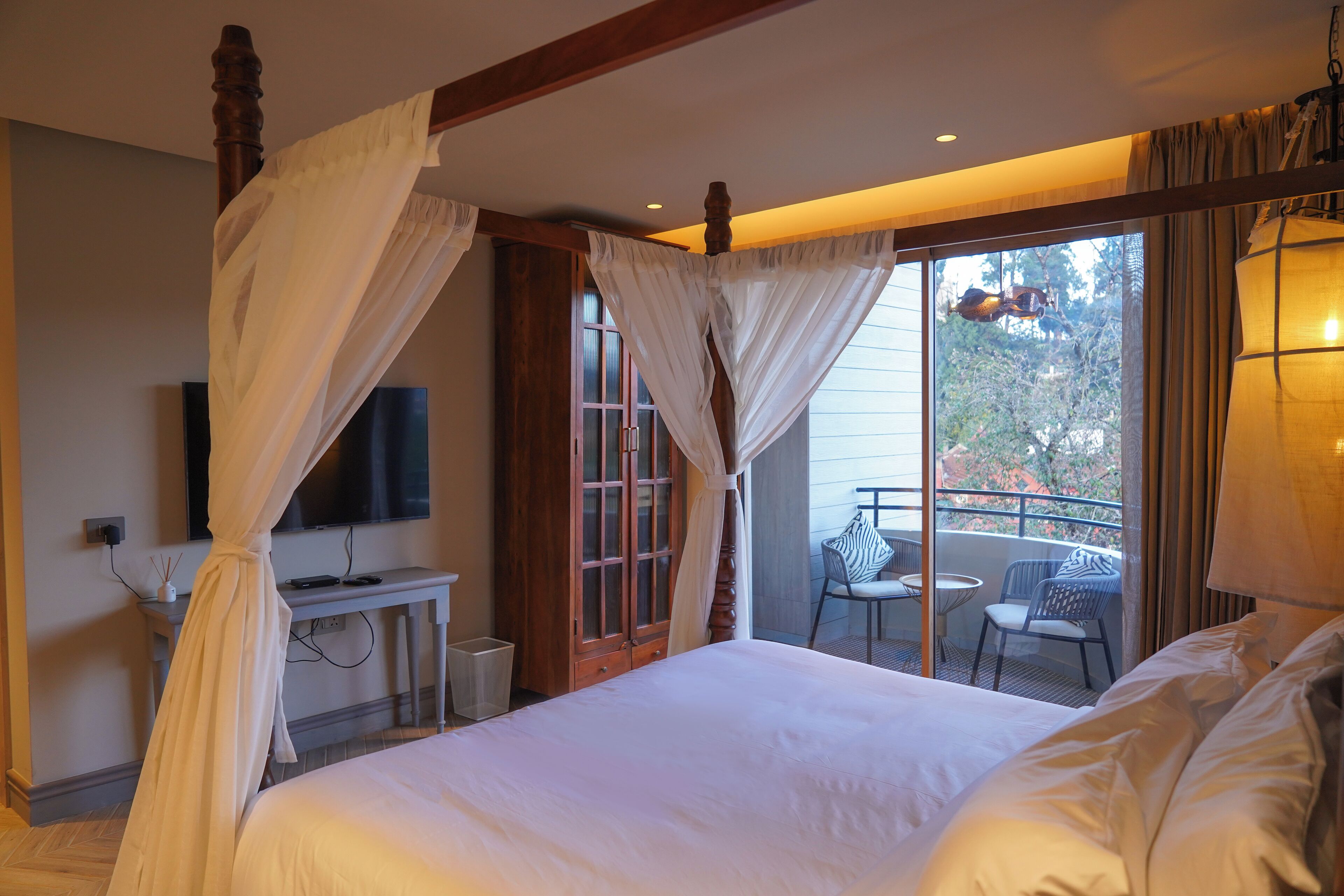 A bedroom with a four-poster bed, a television on the wall, and a balcony with outdoor seating.