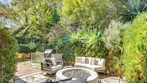 Un patio con una chimenea de piedra, mobiliario exterior y abundante vegetación.