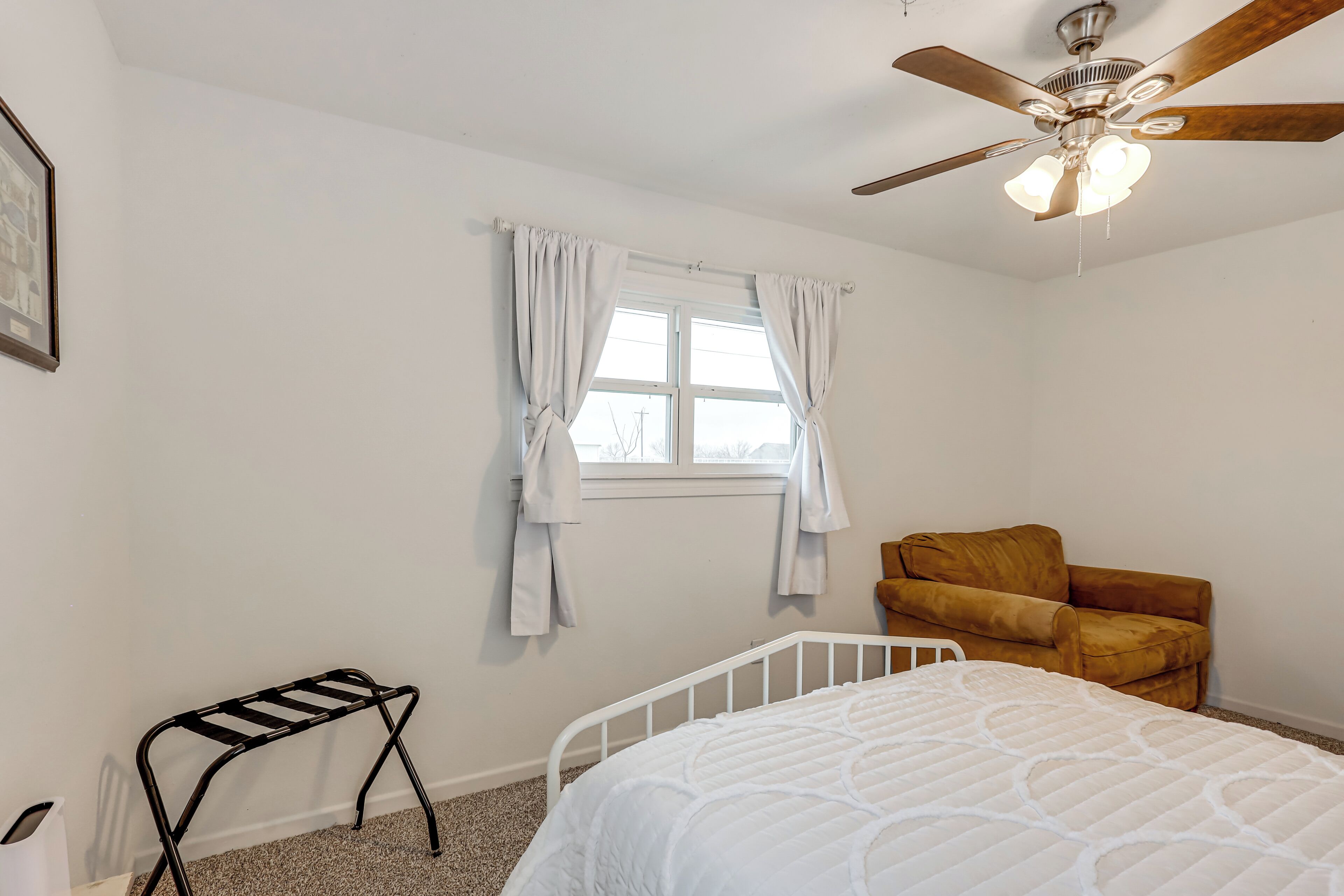 A bedroom with a white bed, a brown chair, a window with white curtains, and a ceiling fan.