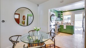 A dining area with a glass table and chairs, a round mirror on the wall, and a view into the kitchen with green cabinets.