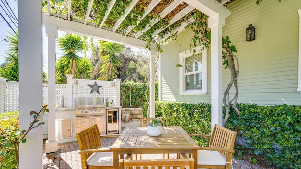 A patio with a wooden table and chairs, a grill, and a pergola with climbing plants.