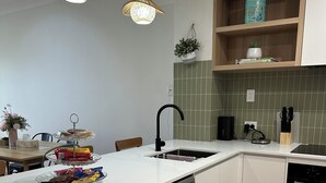 Een moderne keuken met witte kasten, een vaatwasser, een spoelbak en een aanrecht met een afdruiprek. Er zijn twee hanglampen, een potplant en een plank met een bak.