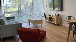 Een moderne woonkamer met een bank, een rode poef, een klein tafeltje en een aan de muur bevestigde televisie.