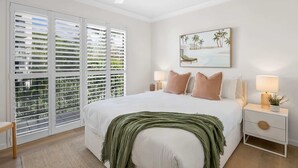 A bedroom with a bed, two bedside tables with lamps, a ceiling fan, a large window with blinds, and a wall-mounted painting. - Bright 2-Bed Apartment with Stunning Ocean Views (Kings Beach)