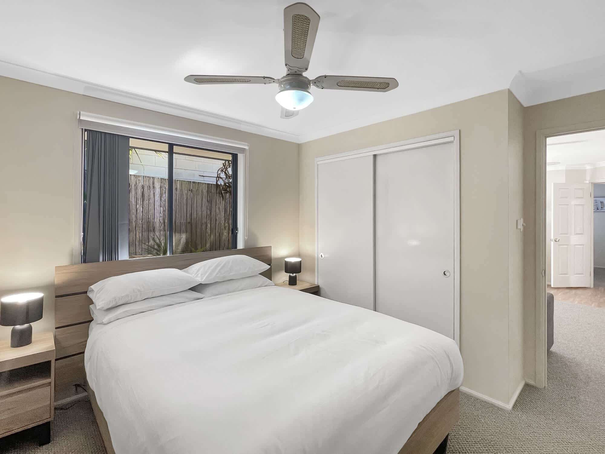 Un dormitorio con cama, lámparas de noche, ventilador de techo, ventana con cortinas y armario.