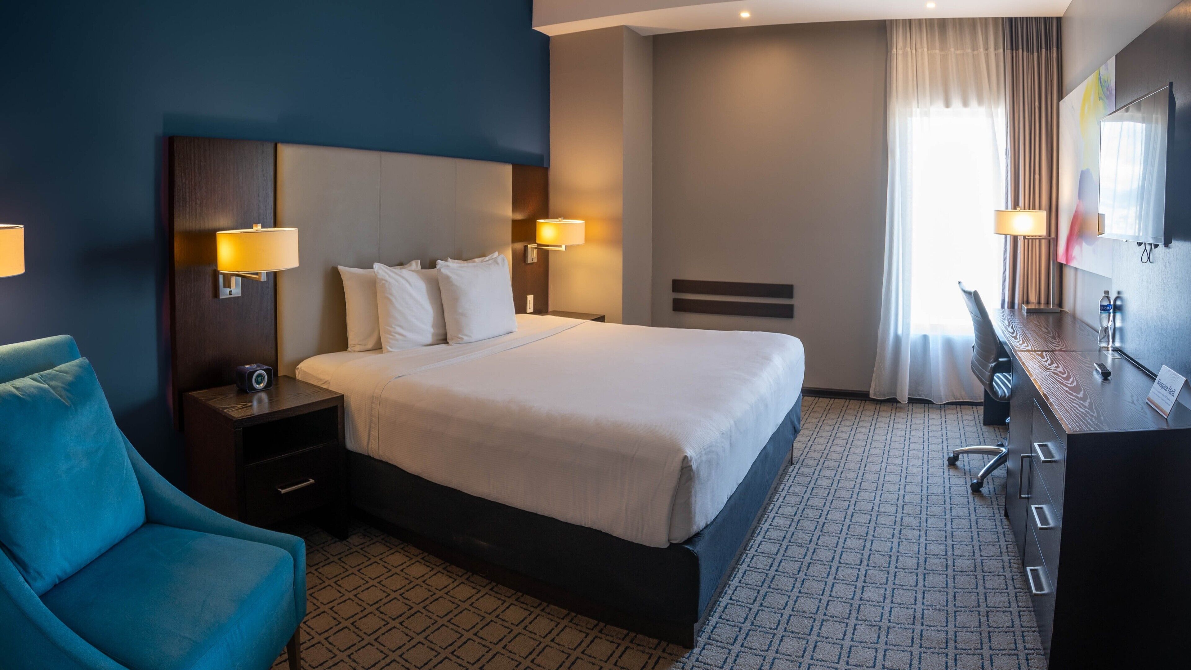A hotel room with a large bed, two bedside tables with lamps, a desk with a chair, a blue armchair, and a large window with curtains.